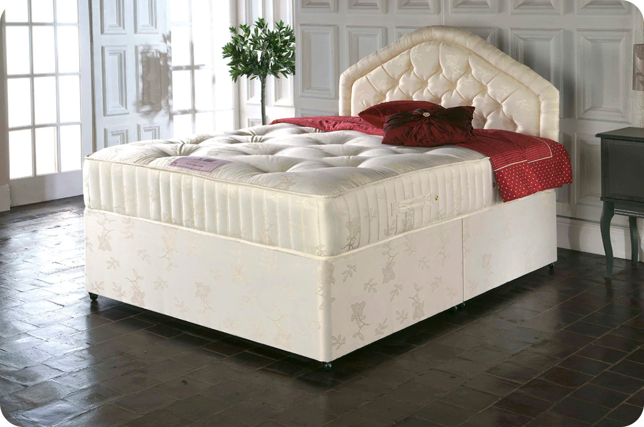 Quilted white mattress on upholstered bed frame with red throw pillows and floral-patterned base