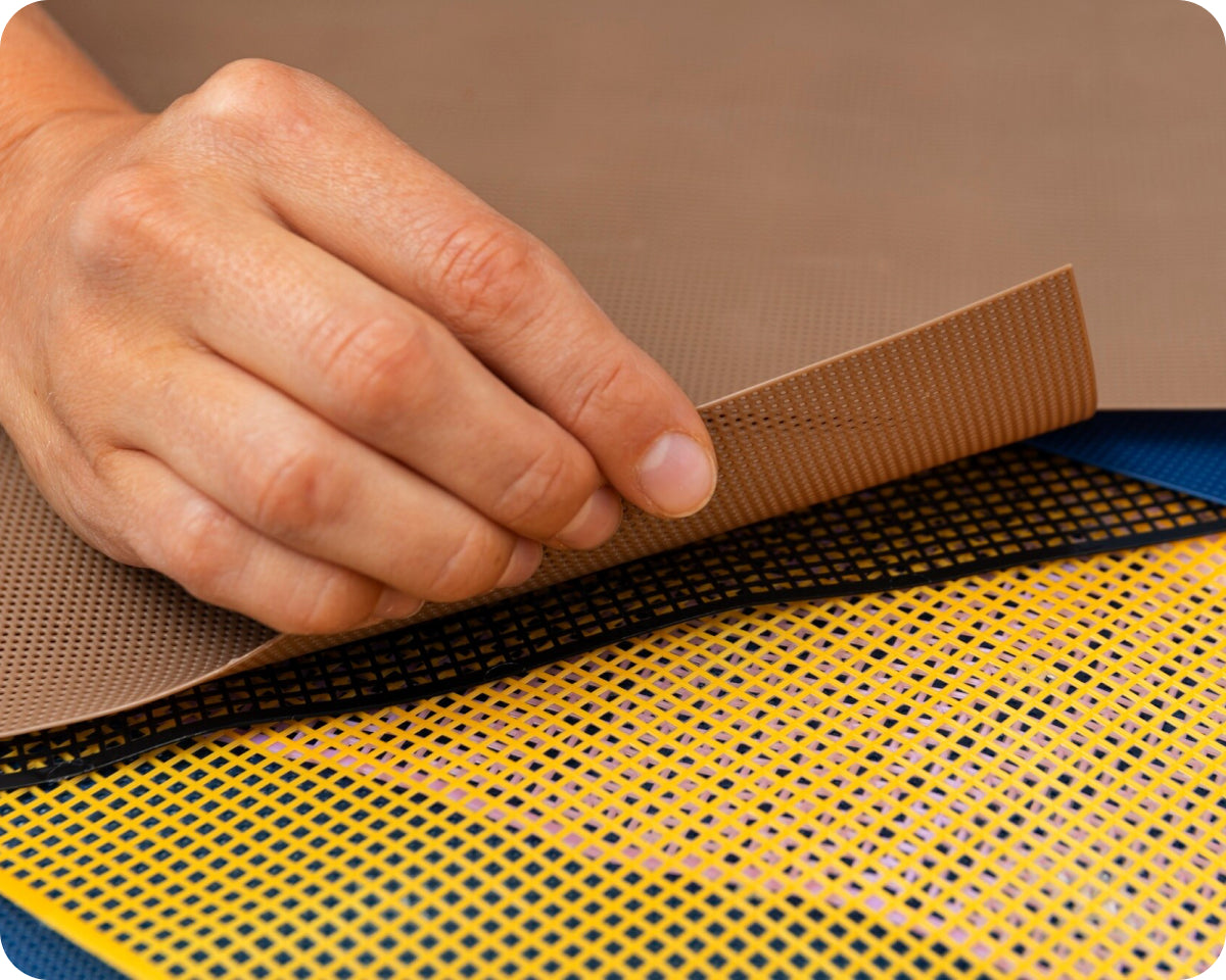 Brown textured silicone mat with yellow mesh backing, hand demonstrating flexibility.
