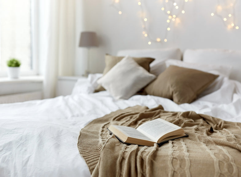 Cozy bedroom with a white bedspread, decorative pillows, and an open book on a brown knit throw.